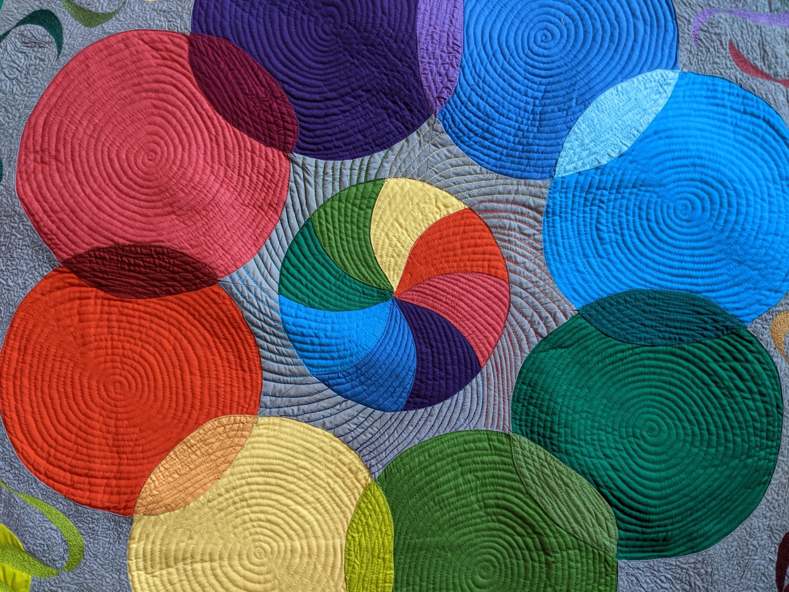 A rainbow swirled circle sewn onto a gray background fabric. The center circle is surrounded by eight larger overlapping circles, each a different color. The whole thing is quilted with swirls and spirals.