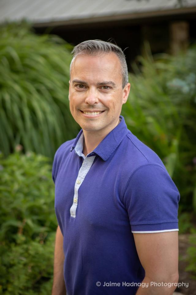 Middle-aged Caucasian man with short salt-and-pepper hair wearing a purple short-sleeved polo shirt and smiling.