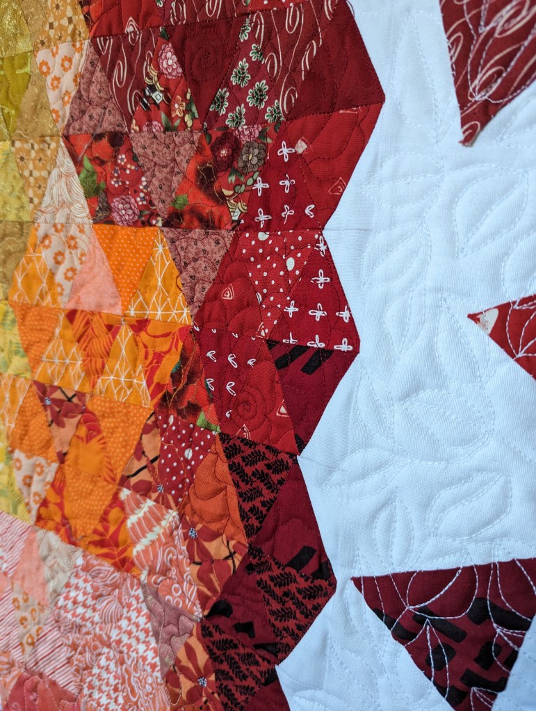 Red, orange, and yellow fabric triangles sewn together on a white background. Leaves are quilted into the white fabric.