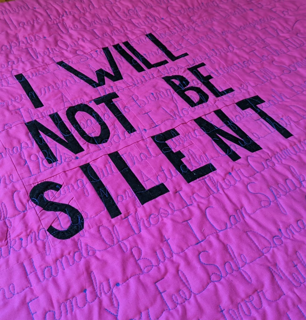 Black fabric words saying "I will not be silent" on a pink fabric background with purple words quilted on top of everything.