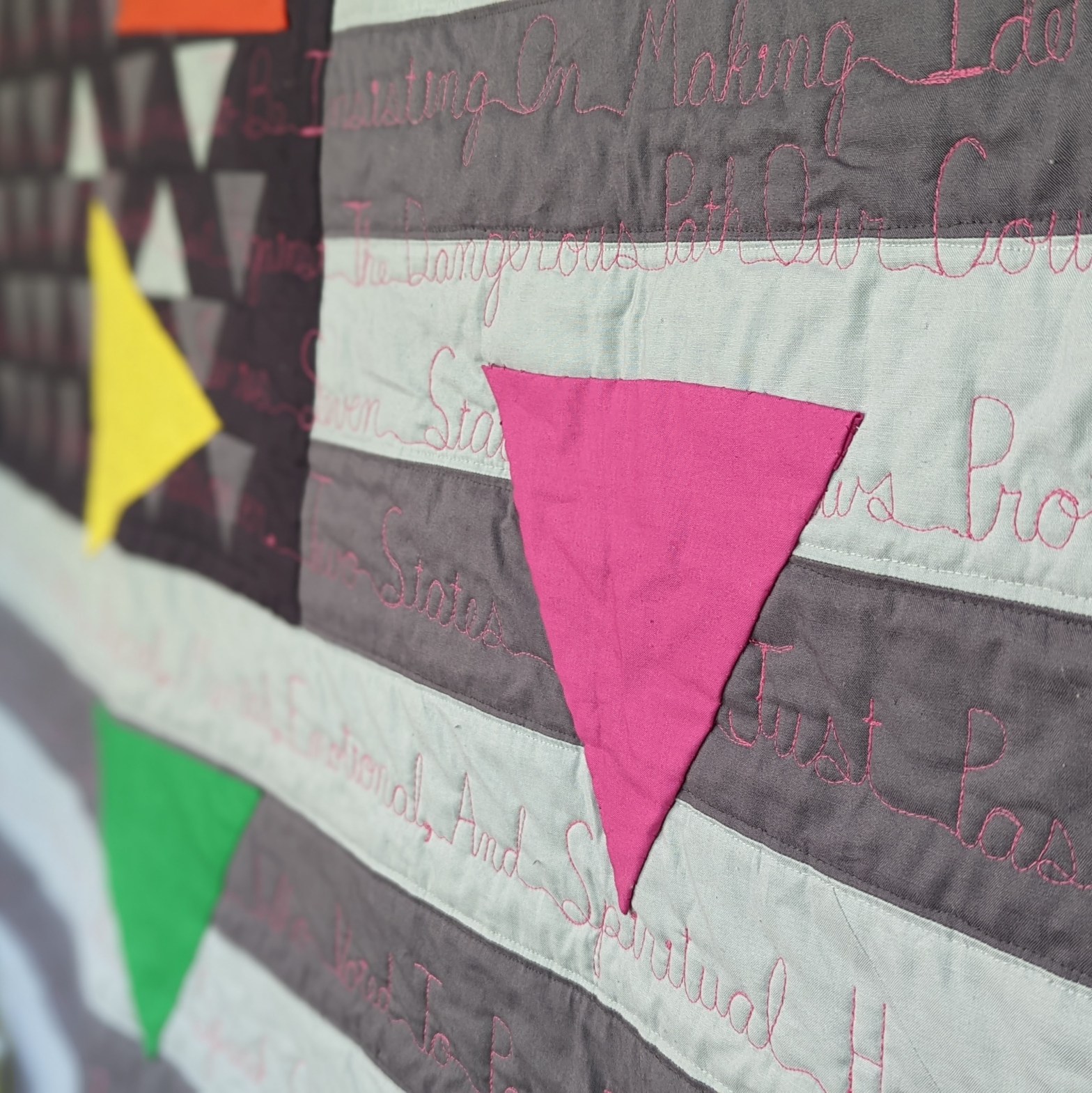 A hot pink triangle sewn onto a gray-scale American flag. A yellow and green triangle are on the left-hand side of the pink triangle.
