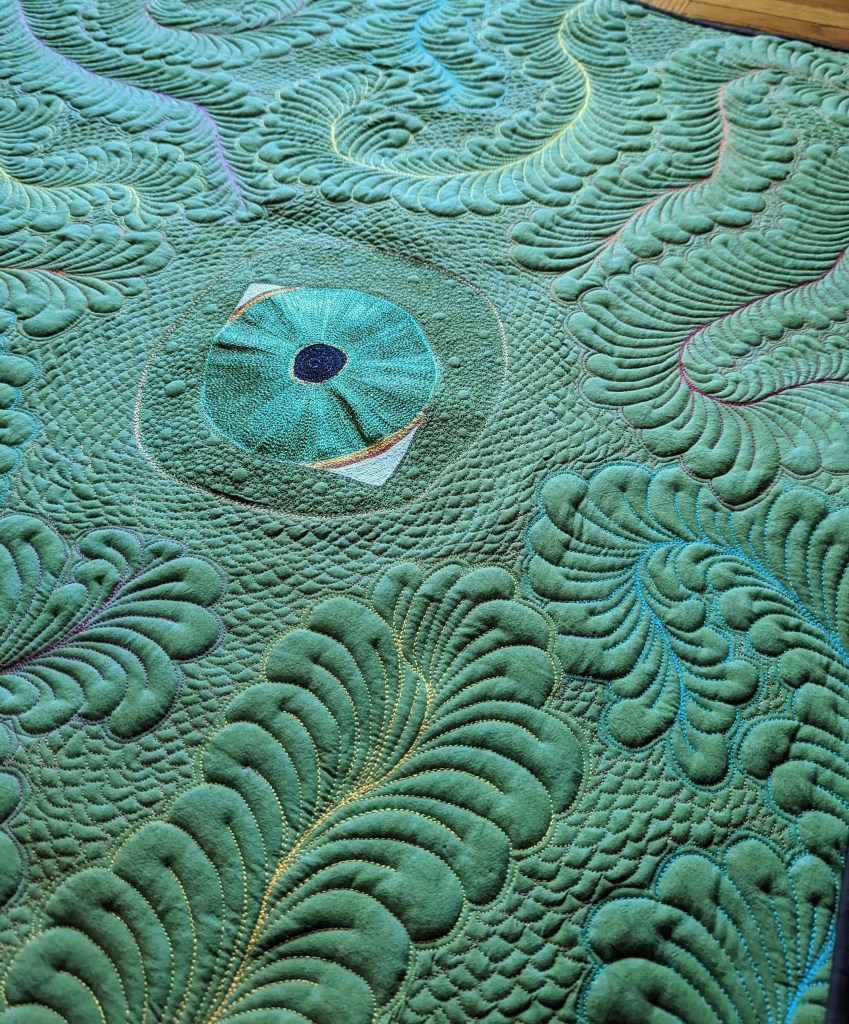 Green fabric that has been heavily quilted to look like a large eye staring through feathers.