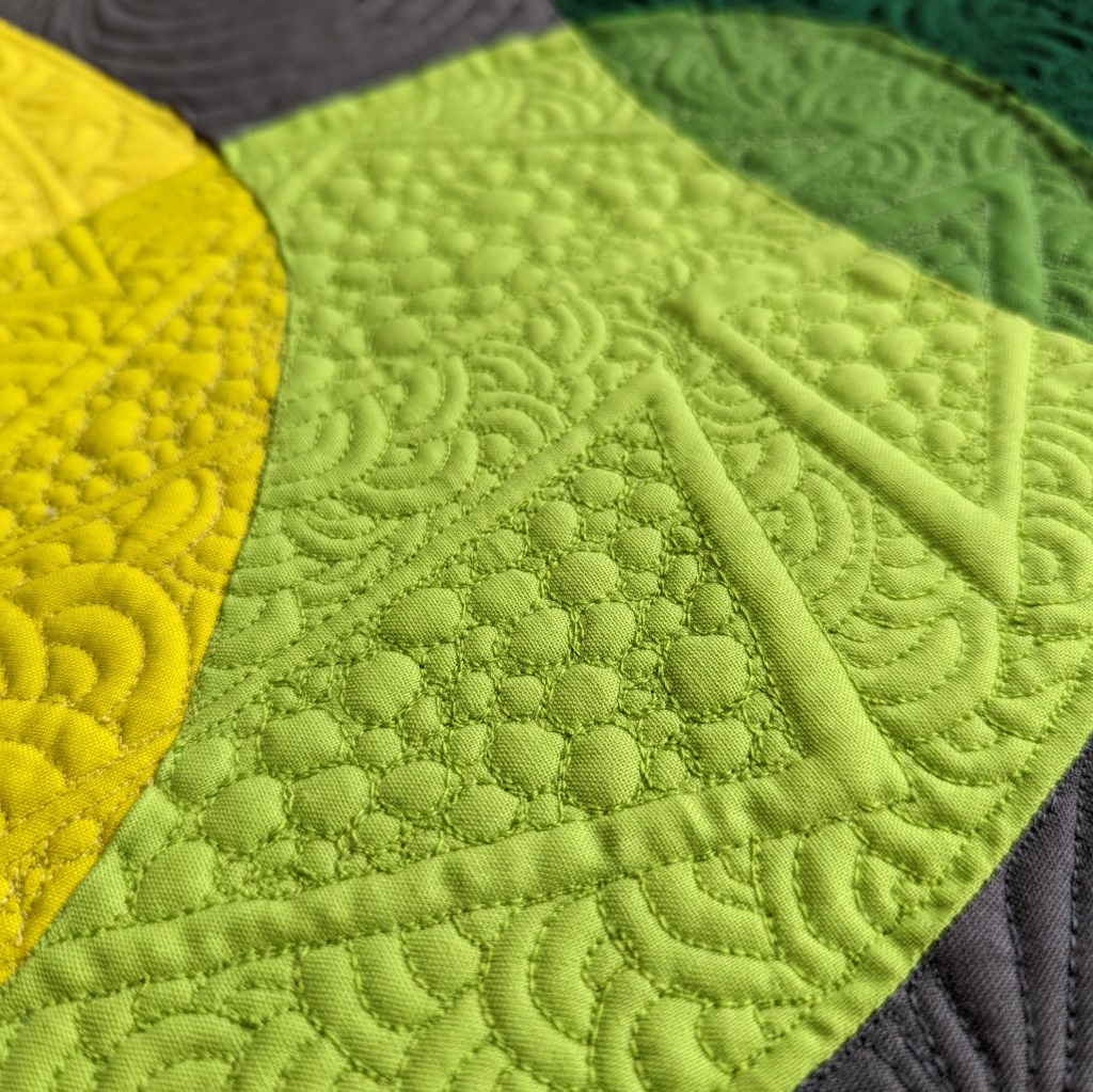 Yellow and green fabric sewn to look like overlapping circle-like shapes that are heavily quilted with swirls and triangles.